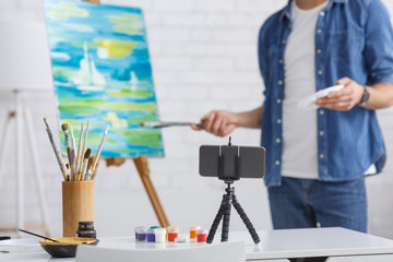 Painting lesson online. Guy painting landscape on easel, on table are glass with brushes and telephone