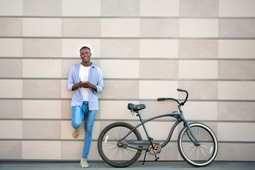 Funky African American guy with bicycle listening to music on smartphone near brick wall