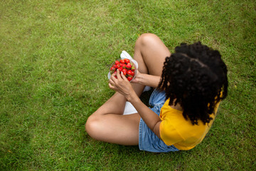 African American girl eating fresh yummy strawberries on green grass at park, top view. Free space