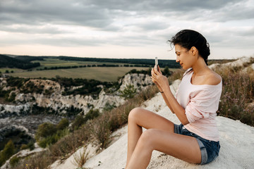 Young woman sitting on a top of the mountain taking pictures of the landscape with smartphone.