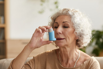 Warm toned head and shoulders portrait portrait of senior woman using inhaler for asthma or breathing problems in home setting - obrazy, fototapety, plakaty