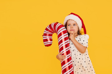 A cute little girl child in a smart dress and a Santa hat holds an inflatable shape of a candy cane on a yellow background. 2021 new year holidays concept and childrens new year gifts advertising