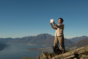 Woman Standing on a Mountain Peak and Taking Photos with Her Tablet over Alpine Lake Maggiore and Mountain in Ticino, Switzerland. © Mats Silvan