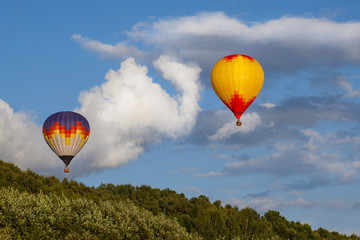 Two multi-colored hot air balloons flying over the summer forest. Some clouds in the sky.