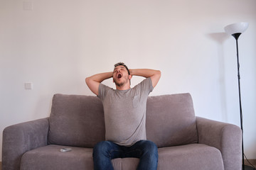 Portrait of a bored and tired young man sitting on a sofa yawning and stretching. Fatigue and tiredness concepts.