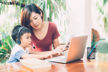 Young asian mother and son using laptop computer and tablet for study and learn together at home, boy wearing headphone for e-learning with distancing, teacher or mom support child, education concept.