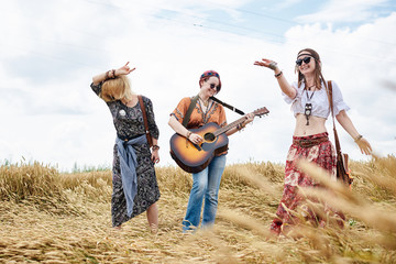 Three hippie women, wearing boho style clothes, dancing in the wheat field, playing guitar, laughing, Female friends, traveling together in countryside. Eco tourism concept. Summer leisure time