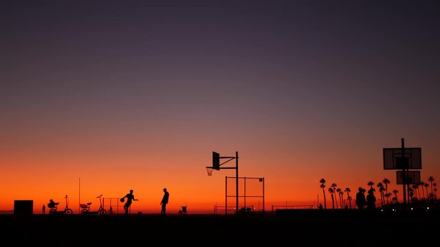 California summertime dusk beach aesthetic, pink sunset. Unrecognizable silhouettes, people play game with ball on basketball court. Newport ocean resort near Los Angeles CA USA. Purple sky gradient.