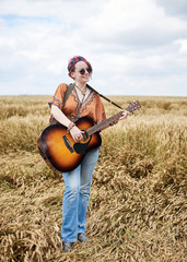 Naklejka na ściany i meble Young hippie women with red hair, wearing boho style clothes, standing on wheat field, playing guitar, smiling. ,Travel musician in countryside. Eco tourism concept. Summer leisure activity.