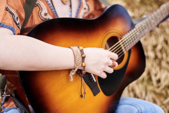 Close-up picture of acoustic guitar held in hands of young hippie women with red hair, wearing boho style clothes, sitting in middle of wheat field,Travel musician in countryside.Musical instrument.