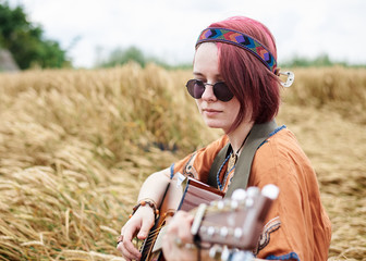 Young hippie women with red hair, wearing boho style clothes, sitting in the middle of wheat field, playing guitar. Travel musician in countryside. Eco tourism concept. Summer leisure activity.