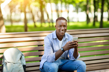 Obraz premium Portrait of African American guy using mobile phone on bench at summer park