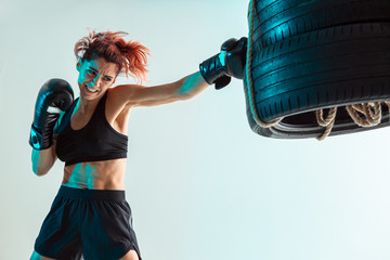 Athletic female fighter boxing on punching bag of tires in studio in neon light. Mixed martial arts...