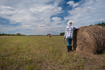 Naklejka na ściany i meble A female farmer stands by a haystack after harvesting in a field with the sky and forest in the background. Landscape