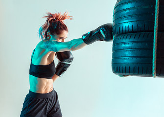Athletic female fighter boxing on punching bag of tires in studio in neon light. Mixed martial arts...