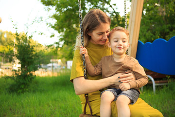 Smiling baby boy and mother ride on swing in playground on green lawn. Summertime photo for ad or...