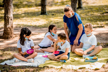 School children in white t shirt eating lunch with teacher during their lunch break at school. They are wearing a protective face masks down.