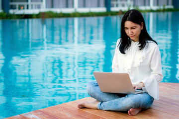 Happy young gorgeous Asian businesswoman in white shirt and jeans working on a laptop by the swimming pool to initiate the idea of creativity and work from anywhere.