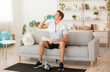 Stay home during quarantine. Guy with towel on shoulders drinks water from bottle, sits on couch with dumbbells