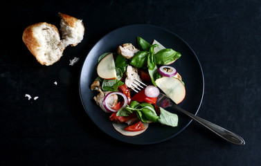 Fototapeta na wymiar Salad with tomatoes, apple, onion, basil and chicken, next to a piece of bread. Dark style