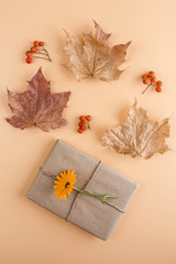 Autumn flat lay composition. Pretty gift box wrapped with craft paper and decorated with flower on orange background among dry foliage. Thanksgiving day concept. Copy space. Selective focus