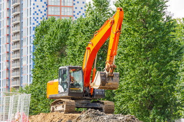 An excavator stands on heaps of soil during the construction of the road against the background of trees.