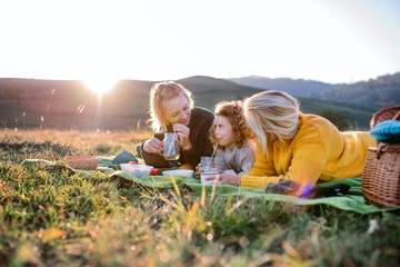 Small girl with mother and grandmother having picnic in nature at sunset.