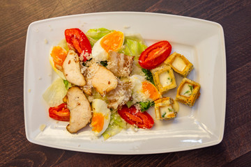 Light, fresh salad with boiled egg, chicken breast, bacon, tomato, lettuce and croutons with sauce in a white rectangular plate on a wooden background. Top view. Healthy food