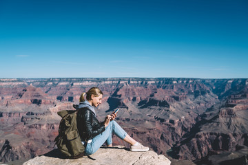 Fototapeta premium Smiling caucasian female travel blogger with backpack using connection on mobile phone for blogging during wanderlust tour in mountains,millennial woman explorer satisfied with connection on cellular