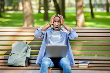 Big failure. Young black guy holding his head in panic near laptop computer at park