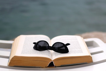 Open book and fashionable sunglasses on a deck chair by the water. Selective focus.