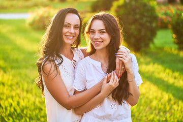 Portrait of stylish attractive smiling joyful happy mother with daughter embracing each other in a park outdoors at sunset at soft light