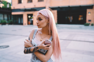 Beautiful Caucasian millennial with tattooed hand holding modern smartphone device for blogging and thoughtful looking away,pink haired woman with cellular technology standing at urbanity and thinking