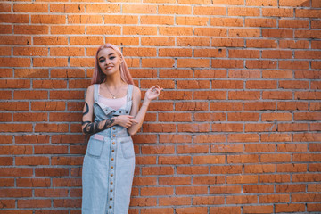 Half length portrait of trendy adolescent teenager dressed in casual jeans sundress smiling at camera near brick promotional background,happy Caucasian hipster girl posing at copy space publicity area