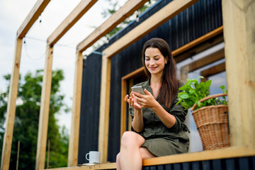 Young woman with smartphone outdoors, weekend away in container house in countryside.