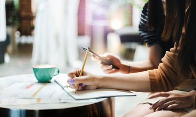 Fototapeta premium Close-up of hand young asian businesswoman holding pencil writing on book while she working with friend and her friend using smart phone searching data from internet