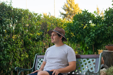 Photo of a young and attractive man in his 20s sitting on a bench enjoying a sunny day. Wearing a grey T shirt and a brown hat. Garden. Flowers. Relaxing day. Holidays. 