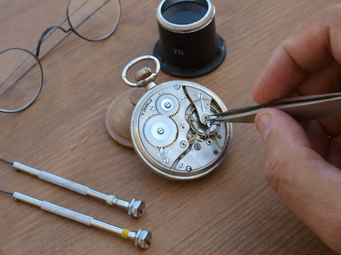 vintage pocket watch under repair, with exposed mechanism, eye glasses and magnifier over wood table