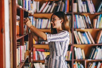 Young gorgeous female student standing next to book shelves and searching for the book for exams. - obrazy, fototapety, plakaty