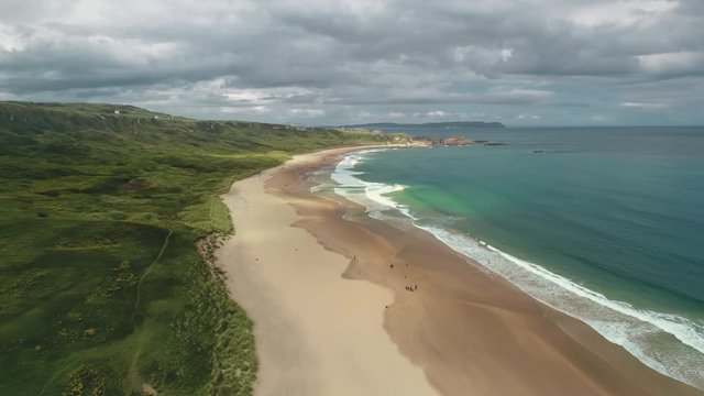 Timelapse Ireland sand beach aerial view: ocean waves, sandy coastline, white shore with greenery meadows. Epic Irish landscape with gray clouds on sky in summer day. Footage cinematic shot in 4K, UHD