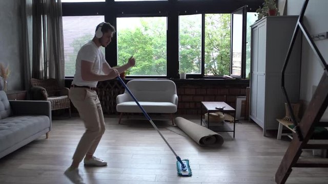 Joyful guy in wireless headphones is singing in mop washing floor at home having fun listening to rock music. Happy people, housework and gadgets concept.