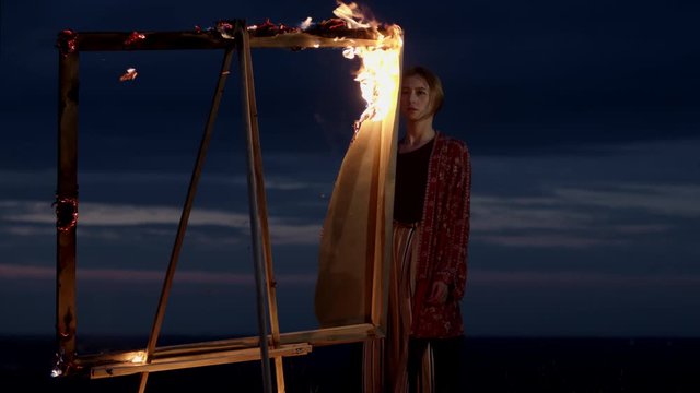 Young Girl Artist Stands In Nature At Dusk In Front Of An Easel With Burning Picture.