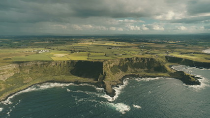 Cliff ocean Ireland coast aerial view: green grass meadows with little cottages in farmland. Amazing autumn rural Irish at deep blue water under grey cloud sky. Cinematic drone shot