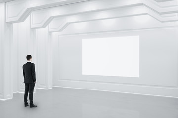 Businessman standing in white classical interior with blank billboard on wall.