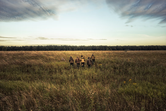 Group of hunters going through rural field towards forest against sunrise sky and forest on horizon during hunting season