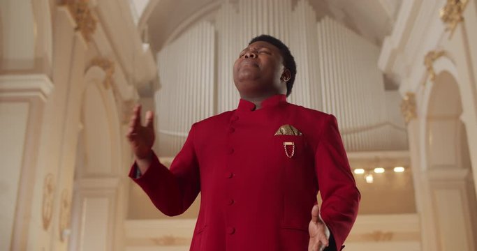 Bottom view of afro american man singing gospel music in church.Portrait of emotional male singer in red suit standing in church and moving hands.Concept of religion and people