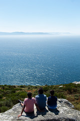 A father with his sons enjoying the breeze and witnessing the Atlantic Ocean while sitting on top of a rock in a viewpoint in the north of Spain