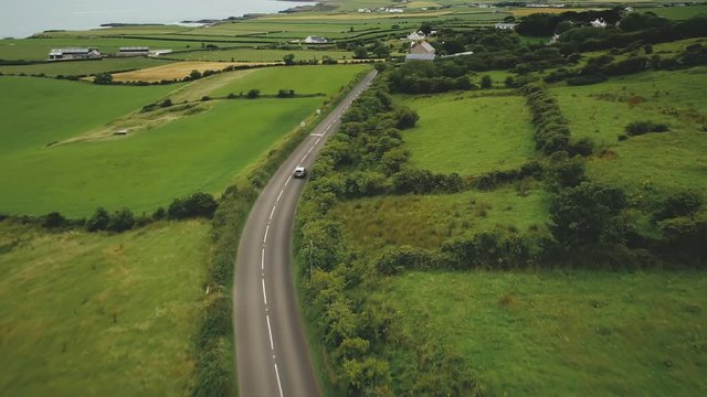 Irish rural road aerial view: green meadows against cottages and hillside farmlands. Picturesque landscape with grain fields in Antrim County, Northern Ireland. Cinematic footage shot in FullHD