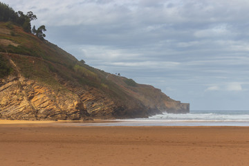 Scenic seascape with mountain in sunlight. Panoramic view of ocean beach and rocks. Cloudy sky above wide empty beach and mountain with cliffs. Calm seaside in bay of Biscay. Travel in Spain. 