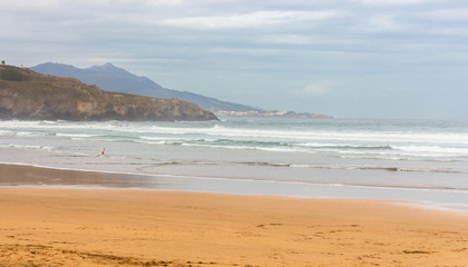 Scenic seascape with mountains and surfer. Cloudy sky above wide beach and mountain with big waves. Seaside in bay of Biscay. Surf concept. Panoramic view of ocean beach and rocks. Travel in Spain. 